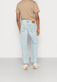 Light blue denim jeans with a straight-leg design, branded leather patch on the back, and slight distressing details. Worn with a beige t-shirt.