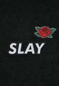 Juodas audinys su žodžiu "SLAY" storais baltais raidėmis, papildytu siuvinėta raudona rože su žaliais lapais.