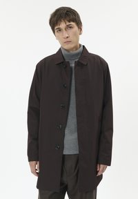 Brązowy długi płaszcz z zapięciem na guziki, z kołnierzem, bocznymi kieszeniami i gładką teksturą, noszony na szarym golfie i brązowych spodniach.