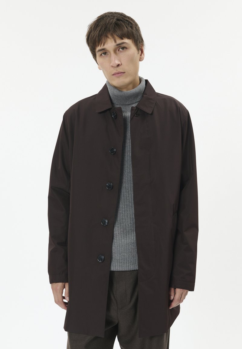 Brązowy długi płaszcz z zapięciem na guziki, z kołnierzem, bocznymi kieszeniami i gładką teksturą, noszony na szarym golfie i brązowych spodniach.