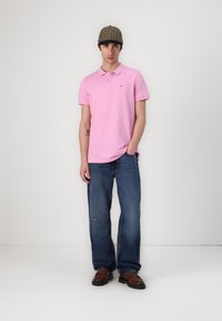 Rosa polo skjorta med en liten logotyp, kombinerad med blåjeans. Modellen bär en mönstrad keps och bruna skor med kraftig sula.
