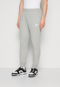 Grå bomullssweatpants med elastisk midja, tapered passform och vit Nike-logotyp på vänster lår, kombinerat med svart och vita sneakers.