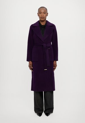 Abrigo largo morado con cintura ceñida, solapas amplias, bolsillos laterales y una textura suave. Combinado con pantalones oscuros de pierna ancha y zapatos de punta.