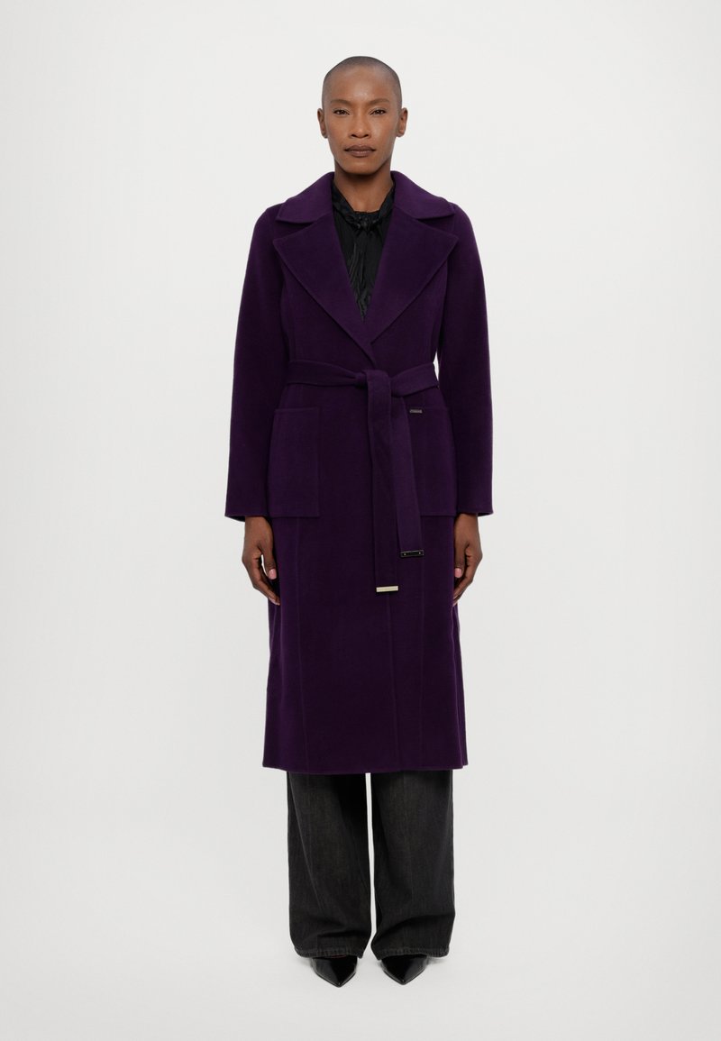 Abrigo largo morado con cintura ceñida, solapas amplias, bolsillos laterales y una textura suave. Combinado con pantalones oscuros de pierna ancha y zapatos de punta.