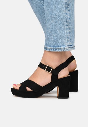 Fødder iført lyseblå jeans og sorte blokhælede sandaler i ruskind med ankelrem og guldlås på hvid baggrund.