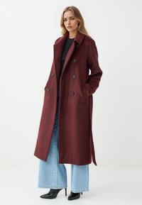 Cappotto bordeaux doppiopetto con colletto, dotato di due tasche laterali e bottoni neri. Il modello indossa jeans azzurri a gamba larga e tacchi.