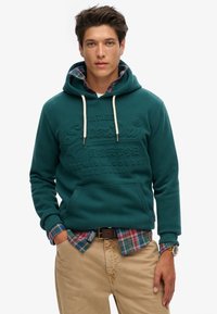 Teal hoodie met trekkoorden, reliëflogo en voorzak, gelaagd over een geruite shirt met blauwe, groene en rode kleuren, gecombineerd met beige broeken.
