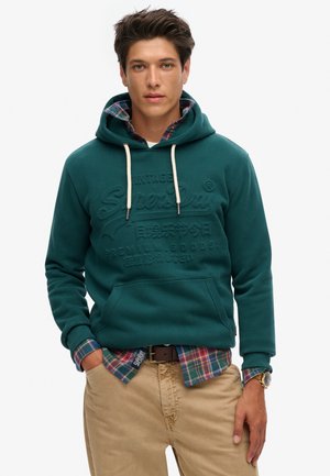 Teal hoodie met trekkoorden, reliëflogo en voorzak, gelaagd over een geruite shirt met blauwe, groene en rode kleuren, gecombineerd met beige broeken.