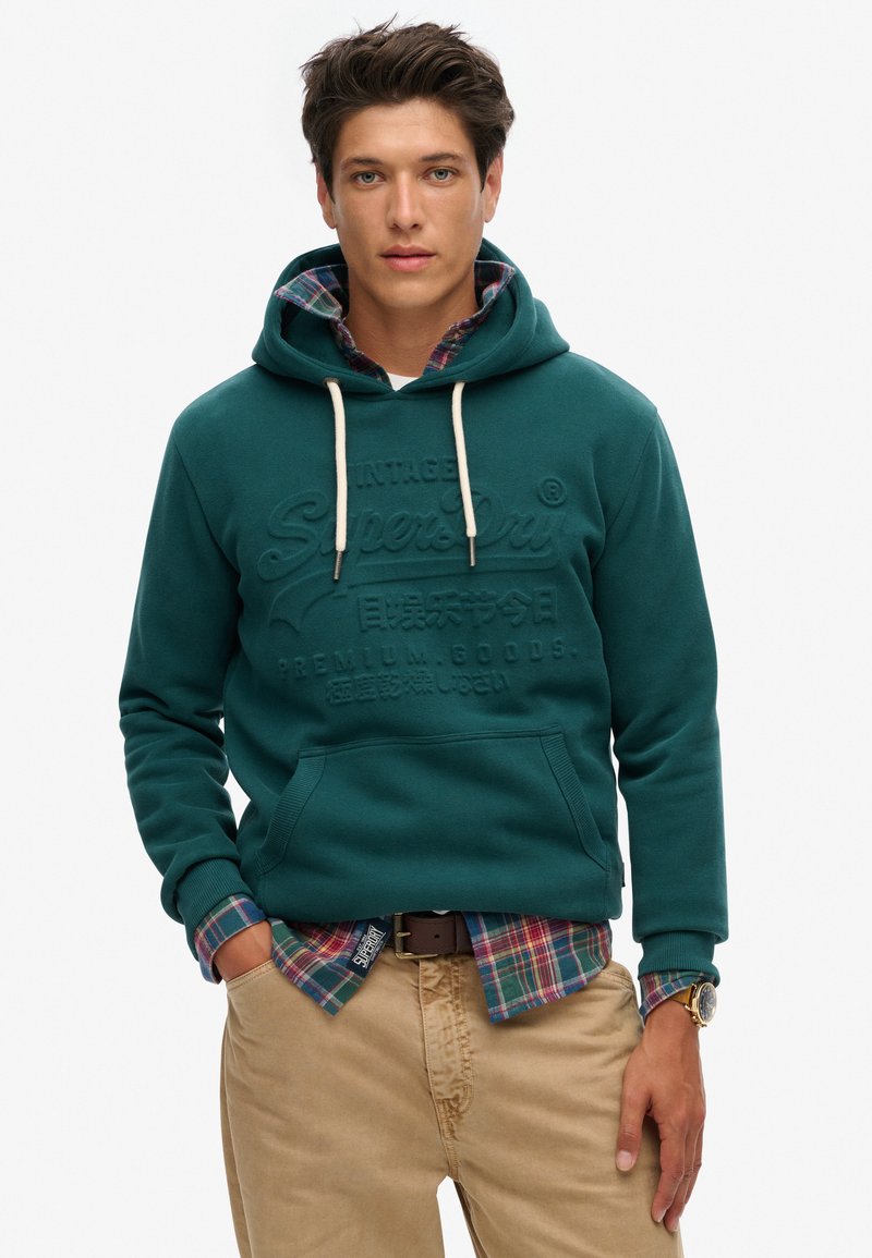 Teal hoodie met trekkoorden, reliëflogo en voorzak, gelaagd over een geruite shirt met blauwe, groene en rode kleuren, gecombineerd met beige broeken.