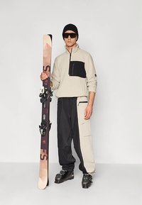 Beige fleece trui met een zwart buitenzakje, zwart-beige skibroek, zwarte skistappers, en ski's met bindingen in de linkerhand, zonnebril op.