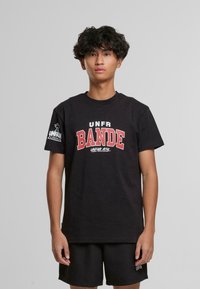 Juodas medvilninis marškinėliai su apvaliu kaklu, ant kurių priekyje yra ryškus raudonas užrašas "UNFR BANDE" ir logotipas ant kairiojo rankovės. Derinami su juodomis šortais.