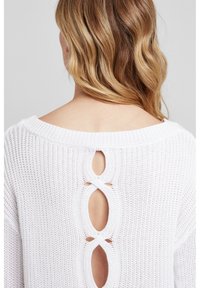 Biały sweter wykonany na szydełku z szerokim dekoltem, otwartym tyłem z dwoma owalnymi wycięciami oraz luźnym, strukturalnym wzorem.