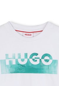 Camiseta blanca con cuello redondo, que presenta un gran diseño gráfico turquesa y la palabra "HUGO" en letras blancas en negrita.