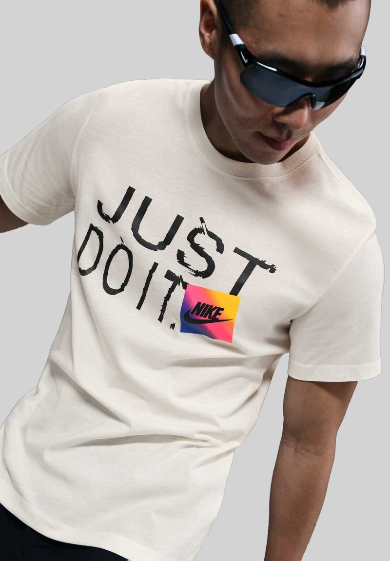 Camiseta color crema con el texto negro "JUST DO IT", que presenta un parche del logotipo de Nike colorido con tonos degradados de rosa y amarillo.