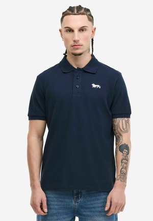 Giovane uomo con capelli intrecciati che indossa una polo blu navy con un logo di un leone bianco e jeans blu, mostrando tatuaggi dettagliati sull'avambraccio destro.