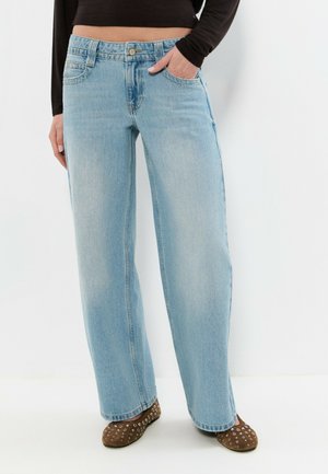 Jeans bleu clair à jambes droites portés avec des ballerines marron à clous et un haut à manches longues noir, une main dans la poche.