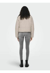 Giacca imbottita corta color beige con una finitura testurizzata, indossata sopra jeans grigi a gamba stretta e stivali neri spessi. Vista da dietro.