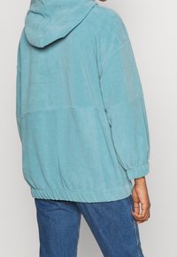 Persona che indossa un felpa leggera blu con cappuccio e polsini ed orlo elasticizzati, abbinata a jeans di denim blu, mostrata di spalle.