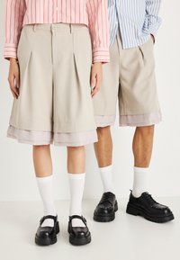 Beežid pleegitud lühikesed püksid, millel on kihiline heledat lillat äärist; mudel kannab musti kingi ja valgeid sokke, mida täiendab roosa ja sinise triibuga särk.