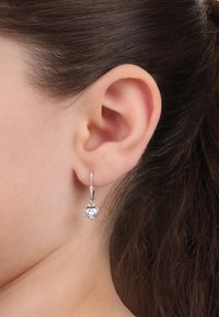 Orecchini a forma di cuore in argento con una pietra trasparente, sospesi a gancio, mostrati su un orecchio con capelli scuri e lisci.