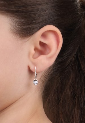 Orecchini a forma di cuore in argento con una pietra trasparente, sospesi a gancio, mostrati su un orecchio con capelli scuri e lisci.
