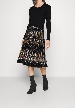 Kvinde iført en sort, langærmet tætsiddende top og et plisseret nederdel i knælængde med flerfarvede geometriske mønstre, kombineret med sorte støvler med hæl.