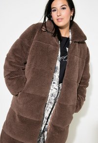 Piumino in pile marrone con colletto alto, caratterizzato da un design trapuntato e una vestibilità ampia, indossato sopra una maglietta stampata con un motivo ben visibile.