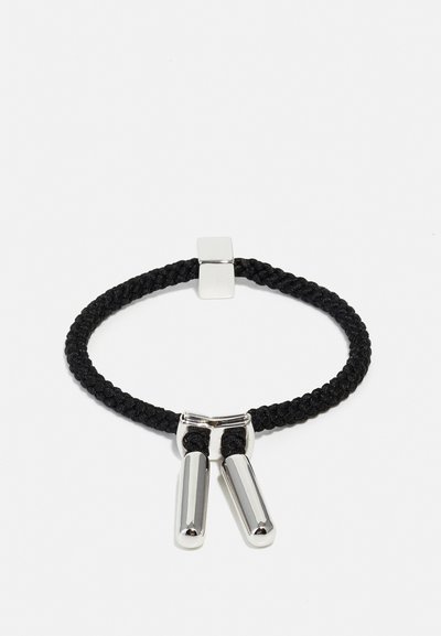 Bracelet tressé noir en textile avec des accents métalliques cylindriques en argent et un fermoir carré. Il présente un design simple et minimaliste.
