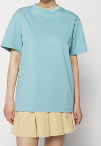 Jasnoniebieski bawełniany t-shirt z krótkimi rękawami, okrągłym dekoltem i logo przy kołnierzu. W zestawie z beżową plisowaną spódnicą.