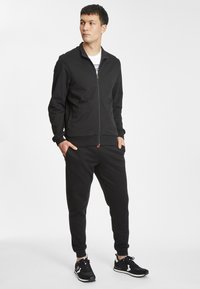 Zwarte zip-up jas en bijpassende joggingbroek, zachte stof, getailleerd ontwerp, geribbelde manchetten, voorzakken, gecombineerd met zwarte sportschoenen.