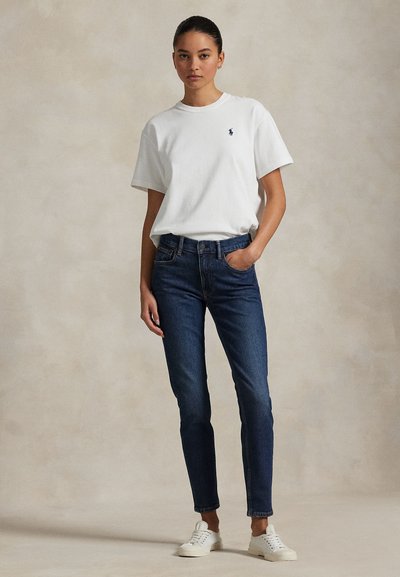 Mladá žena stojí oblečená v bielom tričku s krátkym rukávom s logom, tmavomodrých džínsoch a bielych teniskách na jednoduchom pozadí.