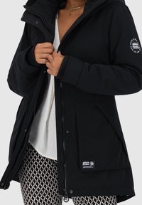 Černá izolační bunda s kapucí, žebrovanými manžetami, předním zipem a logem. Nosí se přes bílý top a vzorované legíny.