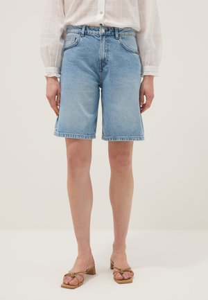 Donna che indossa pantaloncini di jeans azzurri fino al ginocchio, una camicia bianca a maniche lunghe con maniche a sbuffo e sandali beige con tacco basso e fiocchi.