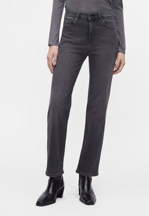 Person iført straight-leg, falmede sorte jeans, sorte ankelstøvler med hæl og en langærmet grå top, stående mod en hvid baggrund.
