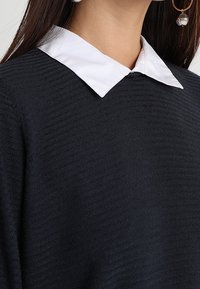 Svart tröja med ett strukturerat stickat mönster, med en vit krage. Nära vy visar tygets ribbade mönster och krags detaljer.