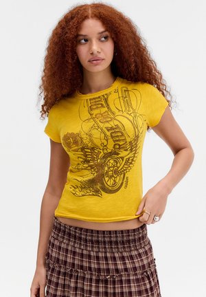 Młoda kobieta z kręconymi rudymi włosami, ubrana w żółty t-shirt z nadrukiem i brązową kratowaną spódnicę, patrzy w lewo, z jedną ręką na biodrze.