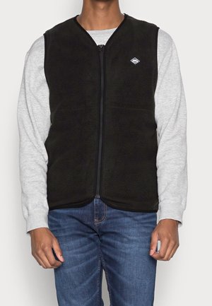 Mann trägt eine schwarze Fleecweste mit Reißverschluss über einem hellgrauen Sweatshirt und blauen Jeans, steht vor einem schlichten Hintergrund.