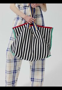 Personne en pantalon à carreaux tenant un grand sac cabas noir et blanc à rayures verticales avec des bords rouges et verts et des poignées bleues et jaunes.