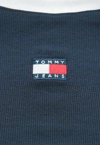Tmavomodrý rebrovaný textil s bielym golierom, s obdĺžnikovou vyšívanou nálepkou v modrej, červenej a bielej farbe s nápisom "TOMMY JEANS".