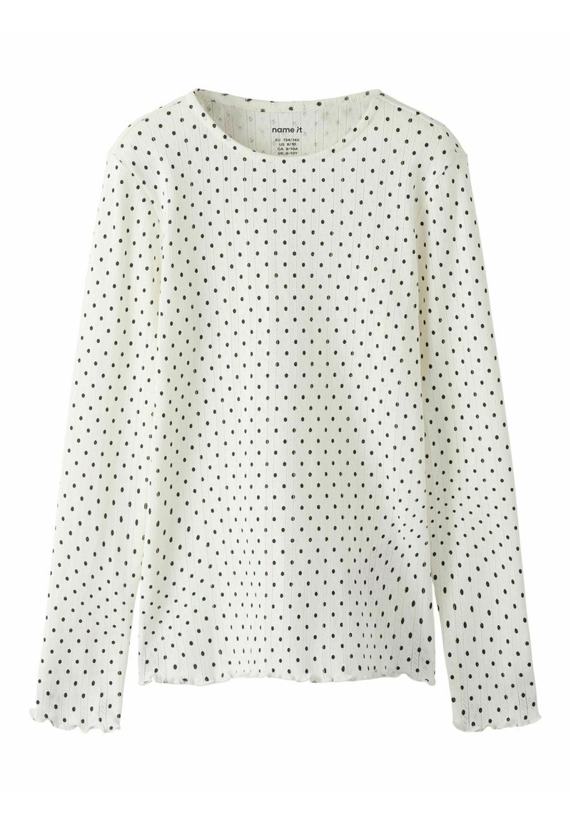 Hvid langærmet top lavet af struktureret stof med sorte polkaprikker. Har rund halsudskæring og bølgede manchetter. Enkel design.