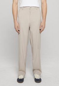 Pantaloni beige chiaro con vestibilità ampia, caratterizzati da una parte anteriore piatta, passanti per cintura e una texture sottile. Abbinati a sneakers blu scuro.