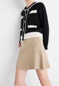 Cardigan nero con bordo bianco e tasche, abbinato a una gonna svasata beige. Entrambi i capi presentano texture morbide e un design classico.