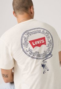 T-shirt en coton couleur crème avec un logo Levi's rouge entouré d'un motif de corde bleue et du texte, accompagné d'un graphisme de cow-boy.