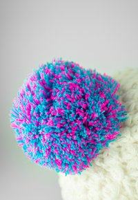 Multicolored pom-pom with blue and pink yarn fibers, fluffy texture, attached to a cream-colored knitted garment or accessory.