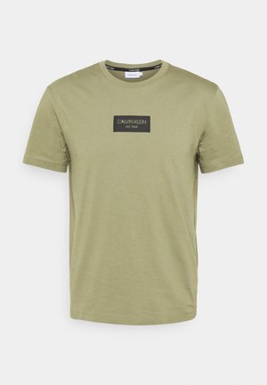 T-shirt à manches courtes vert olive clair avec logo rectangulaire Calvin Klein noir et texte "EST. 1968" sur la poitrine.