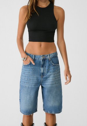 Kvinde iført en sort ærmeløs cropped top og mid-lange blå denimshorts med metalnitter, stående mod en ensfarvet baggrund.