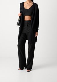 Cárdigan largo negro sobre un top corto negro y pantalones de cintura alta. Tejido liso, ajuste relajado y delantero abierto con bolsillos laterales. Sandalias negras.