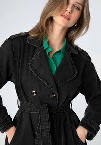 Cappotto di jeans nero con design doppiopetto, bottoni dorati e vita regolabile con cintura. Presenta un ampio colletto e tasche laterali.