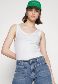 Top de alças branco sem mangas com decote arredondado, combinado com jeans de ganga azul de cintura alta. Modelo usa um boné verde com texto.