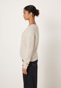 Sieviete ar tumšiem, cirtainiem matiem sasietiem bungā, tērpusies gaiši pelēkā pūkainā džemperī un tumšās biksēs, stāvot profilā pret vienkāršu fonu.